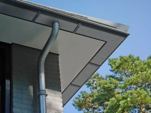 Zinken regenpijp die loopt vanaf een overstek van Trespa tot beneden, met robuuste wrongen.